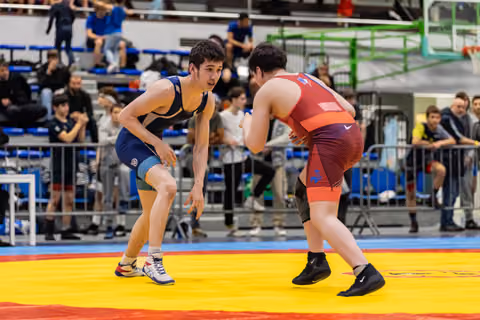 Teen wrestler in blue singlet squares off against opponent in red, both crouched in defensive stance on yellow mat.