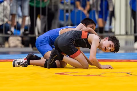 Young wrestler in black singlet controls opponent on yellow mat, focused expression during intense match