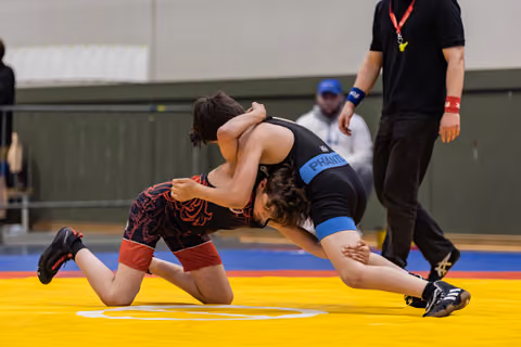 Two young wrestlers locked in a headlock battle on a yellow mat, referee observing closely in the background.