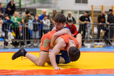 Young wrestler in blue dominates opponent in red, pinning him to the yellow mat during an indoor tournament.