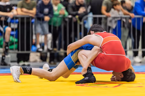 Wrestler in red singlet drives opponent in blue to the mat, executing a powerful takedown during a competitive bout.