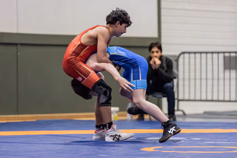 Young wrestler in red drives opponent in blue backward across the mat, both athletes focused and low in a grappling stance.