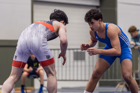 Two young wrestlers in blue and white singlets square off in a tense standoff, eyes locked, bodies coiled ready to engage.