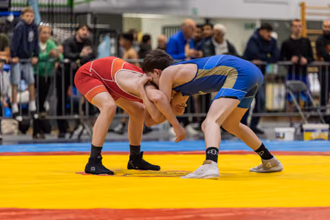 Two young wrestlers in red and blue singlets locked in a clinch, both bent low with intense focus on a competition mat.