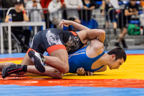 Wrestler in blue singlet being pinned face-down on colorful mat by opponent in black tiger-print singlet.