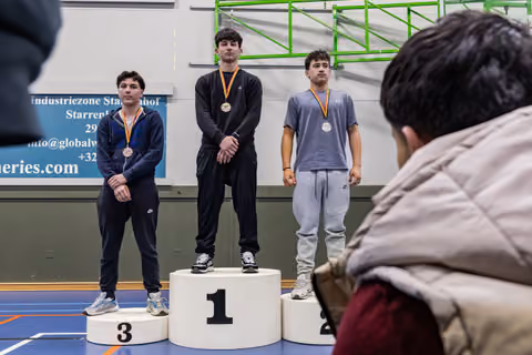 Three young male athletes stand on podium positions 1, 2, and 3, wearing medals, looking solemn in a gymnasium.