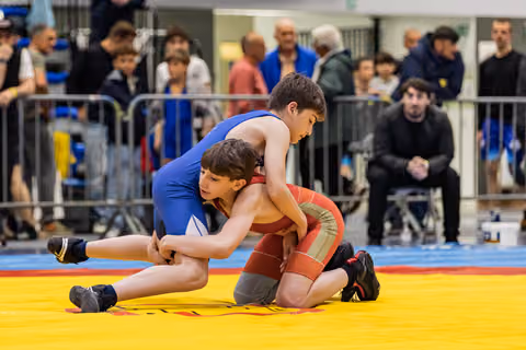 Two boys in blue and red singlets locked in an intense wrestling grapple on a yellow mat, crowd watching behind barriers.