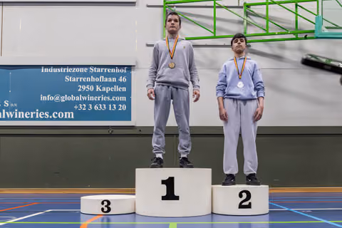 Two young athletes stand on podiums 1 and 2, wearing medals, expressions serious and composed, in an indoor sports hall.