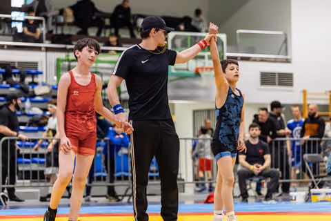 Referee raises the hand of the victorious boy in blue as the defeated boy in red stands nearby, looking dejected.