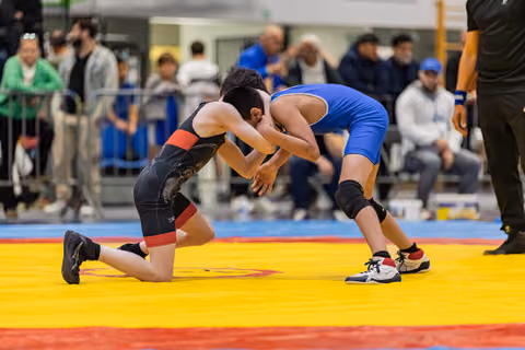 Two young wrestlers in black and blue singlets locked in a match, one on his knee on a yellow and red mat.