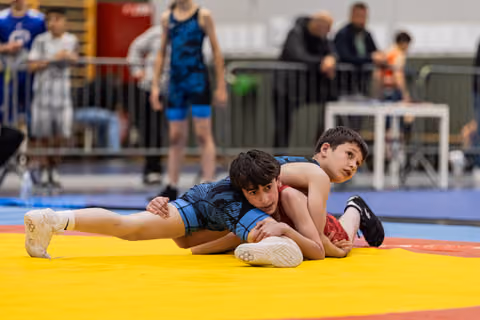 Two young wrestlers on a yellow mat, one in blue controlling his opponent from behind, both focused and determined.