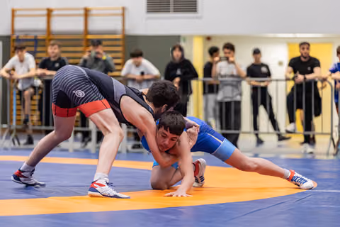 Young wrestler in blue singlet fights to escape a takedown attempt on an orange mat, crowd watching behind barriers.