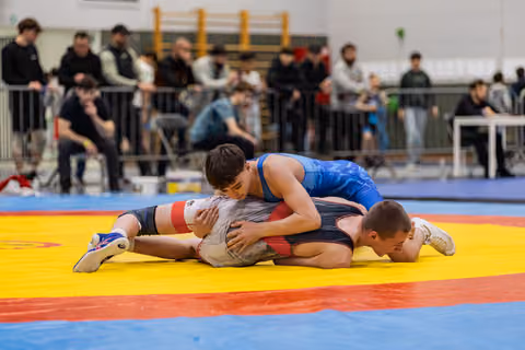 Wrestler in blue singlet pins opponent to yellow mat, showing focused determination during indoor tournament.