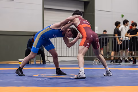Two young wrestlers lock in an intense clinch on the mat, the athlete in blue straining against his Belgian opponent in maroon.