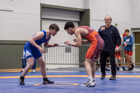 Two young wrestlers square off in intense stance on blue mat, referee watches closely in background.