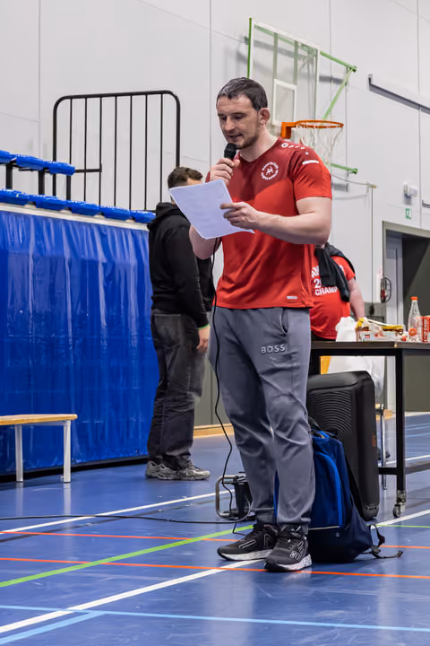 Muscular man in red sports shirt reads from paper while speaking into microphone in indoor sports hall.