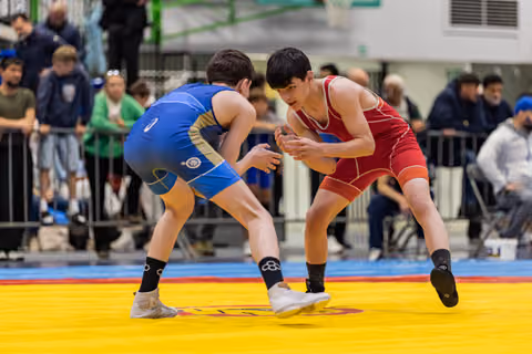 Young wrestler in red singlet squares off against opponent in blue, both in aggressive stance on yellow competition mat.