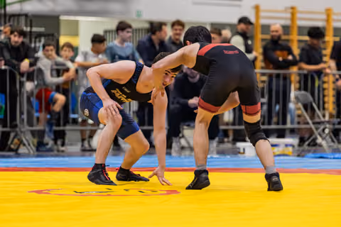 Two young wrestlers in a tense standoff, crouched low on a yellow mat, spectators watching closely in the background.