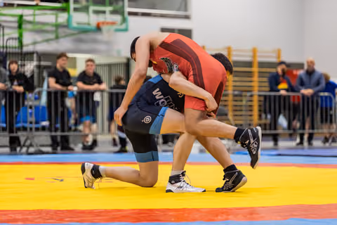 Two wrestlers battle for control, one in blue singlet on knee, other in red driving forward on colorful competition mat.