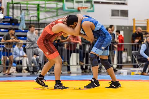 Two wrestlers locked in a clinch on a yellow mat, both hunched forward, straining for control in a busy gymnasium.