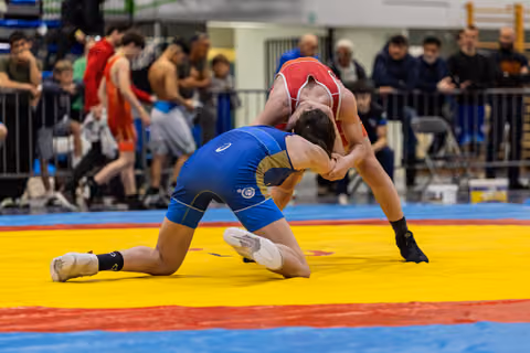 Wrestler in blue singlet on knees, locked in tight clinch with red-clad opponent on colorful mat, intense struggle.