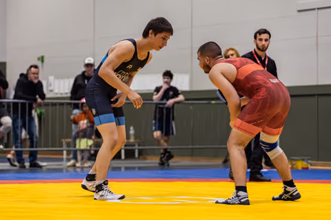 Two wrestlers in blue and red singlets square off in a tense standoff on a yellow competition mat, crowd watching behind.