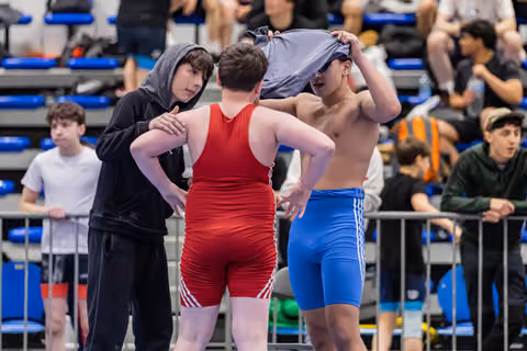 Teen wrestler in blue shorts removes shirt while a teammate in black hoodie offers advice before their bout.