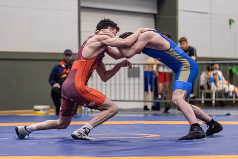Two young wrestlers locked in a tense clinch on a blue mat, both leaning aggressively into each other during a match.