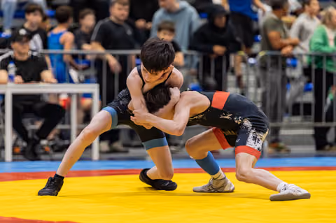 Two young wrestlers locked in a tight clinch, straining with effort on a colorful competition mat before a crowded arena.