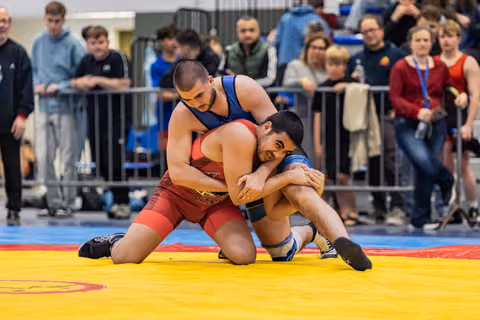 Wrestler in red singlet controls smiling opponent on yellow mat, crowd watching in background.