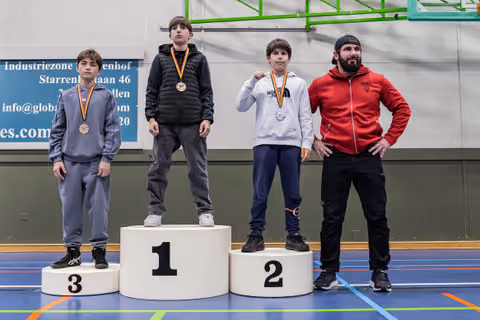 Three young wrestlers stand on a 1-2-3 podium with medals; an adult coach in red stands proudly beside them.