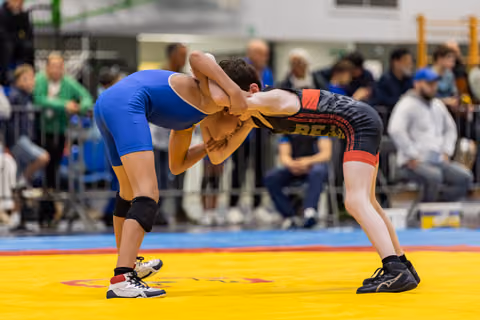 Two young wrestlers in a tight clinch on a yellow mat, one in blue, one in black and red, crowds watching behind.