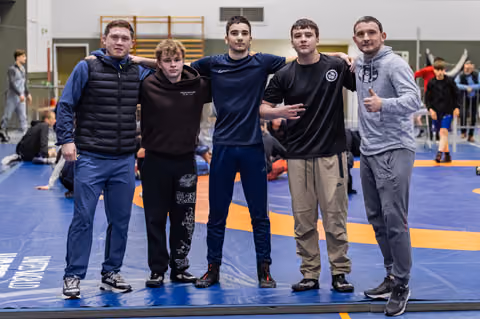 Five young wrestlers pose arm-in-arm on the blue mat, smiling confidently at a training hall event.