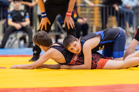 Two young wrestlers grapple on a yellow mat, one pinning the other down, face showing fierce concentration.