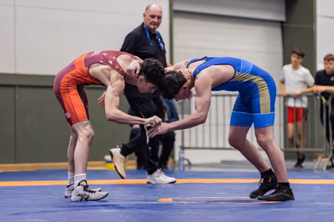Two young wrestlers lock hands in a tense standoff on a blue mat, referee watching closely behind them.