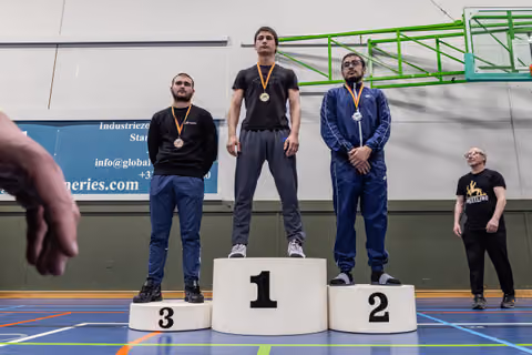 Three male wrestlers stand on a podium, gold medalist centered on platform 1, flanked by silver and bronze winners in a gym.