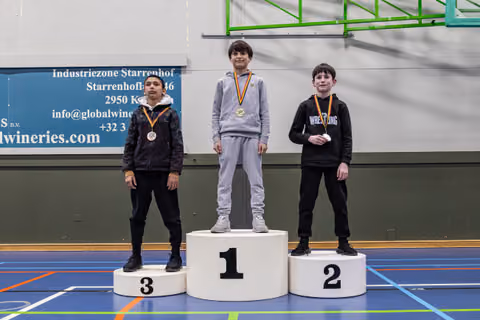 Three young wrestlers on a podium; gold medalist stands tall in center, flanked by bronze and silver winners, all wearing medals.