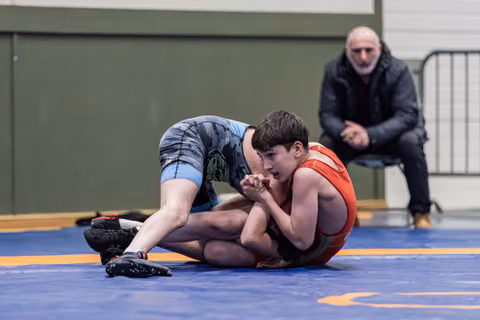 Young wrestler in red singlet holds defensive position on the mat while opponent in blue applies pressure, coach watching.