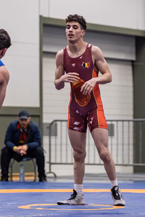 Young Belgian wrestler in maroon singlet stands alert on blue mat, hands ready, focused expression during bout.