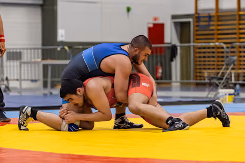Wrestler in blue singlet controls opponent in red, pinning him down on a yellow and red mat indoors.