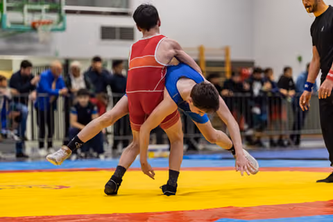 Wrestler in blue singlet being lifted and thrown by red opponent on colorful mat, referee watching nearby.