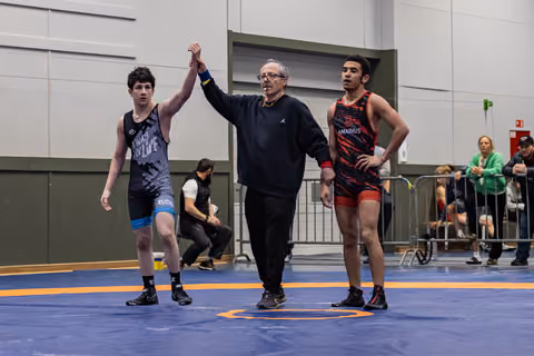 Referee raises the left wrestler's hand in victory as the opponent stands aside, looking composed on the blue mat.