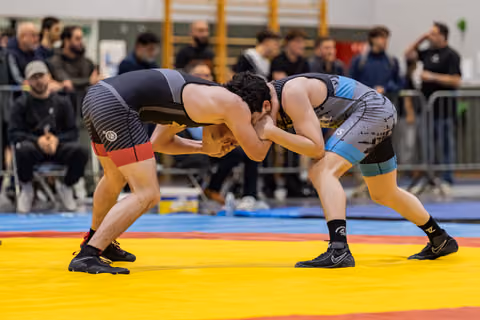 Two young wrestlers lock heads in an intense grappling stance on a yellow and red mat, crowd watching behind.