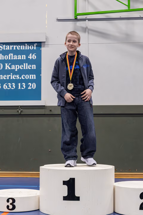 Smiling boy stands proudly on the first-place podium, wearing a gold medal on a tricolor ribbon in a sports hall.
