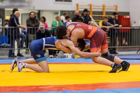 Two wrestlers lock heads in a tight clinch on a yellow mat, blue singlet athlete on one knee fighting for control.