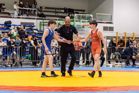 Referee joins hands of two young wrestlers in blue and red singlets before their bout on a colorful mat.