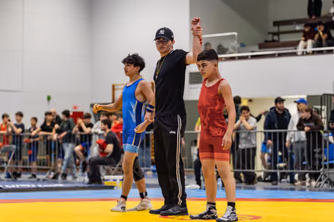 Referee raises the arm of a young wrestler in red singlet declaring victory, while defeated opponent in blue walks away.