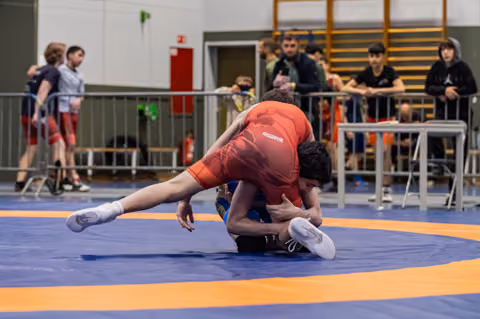 Wrestler in red singlet executes a takedown, pinning opponent to the blue mat as spectators watch from behind barriers.