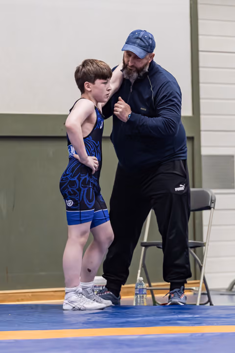 Bearded coach in navy cap leans in to give focused advice to a young wrestler in a blue singlet on the mat.