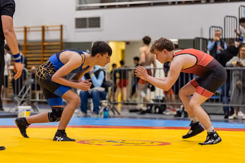 Two teenage wrestlers crouch in tense standoff on yellow mat, hands raised, focused expressions in a gymnasium.